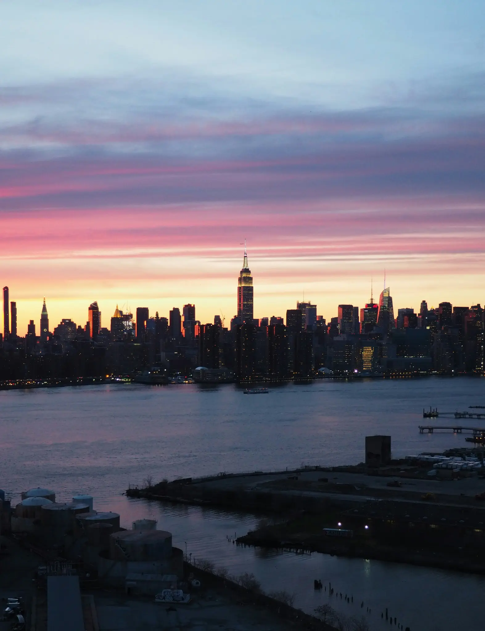 Živý západ slnka nad panorámou mesta s dominantným Empire State Buildingom, pohľad cez tmavú vodnú plochu.
