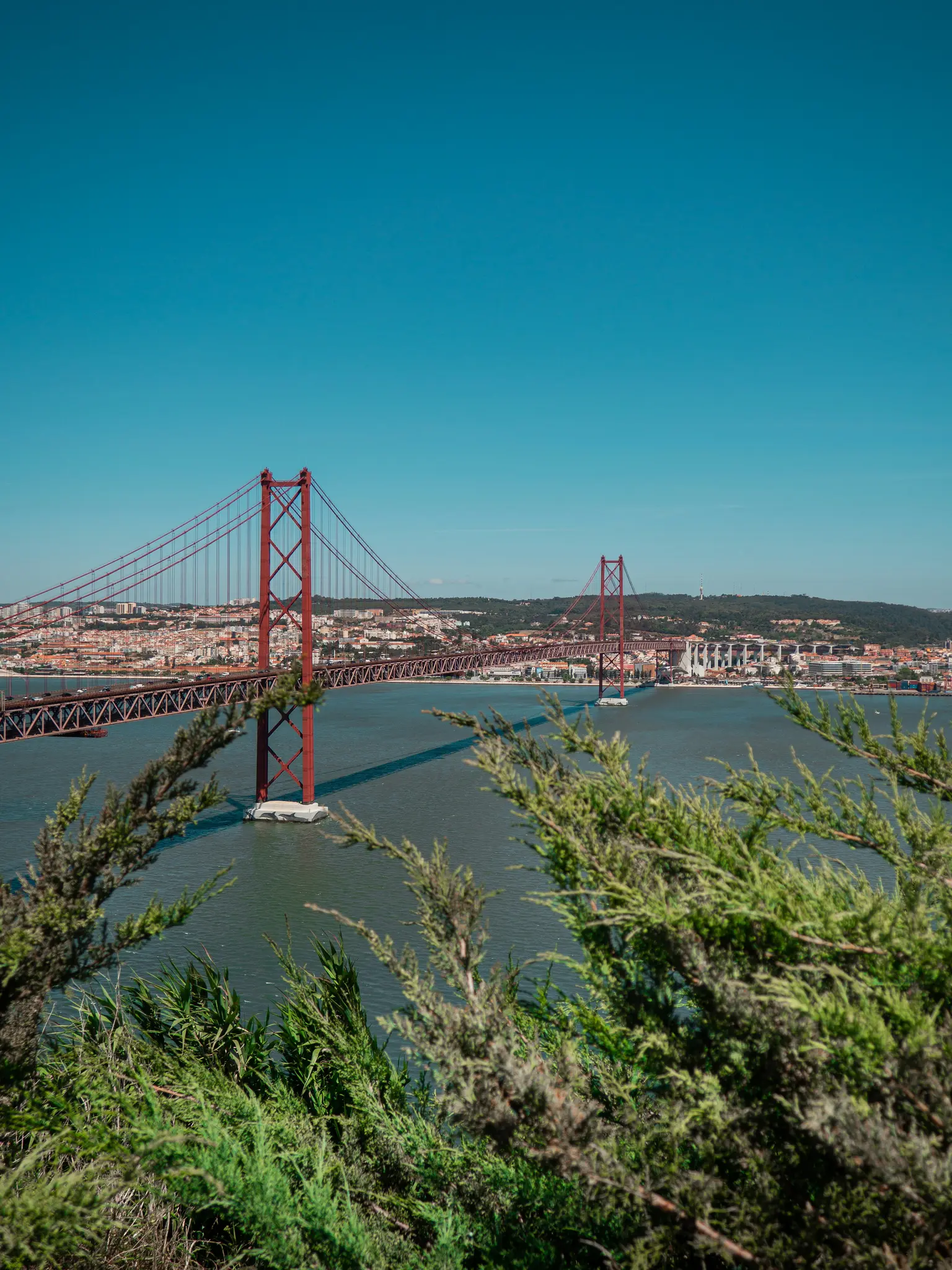 Un puente colgante rojo se extiende sobre una amplia masa de agua bajo un cielo azul claro. Una ciudad bordea la orilla
