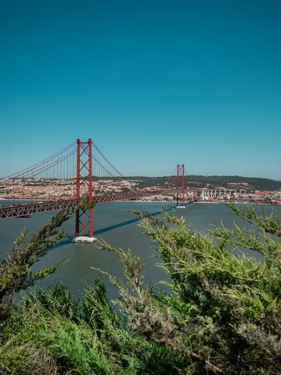 Červený visutý most se klene přes širokou vodní plochu pod jasně modrou oblohou. Na vzdáleném břehu se táhne město.