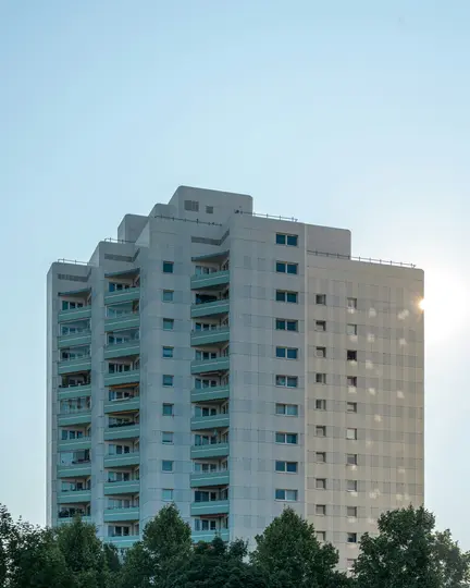 Vysoký, světlý bytový dům se stupňovitou střechou a tyrkysovými balkony na pozadí jasně modré oblohy. Stromy…