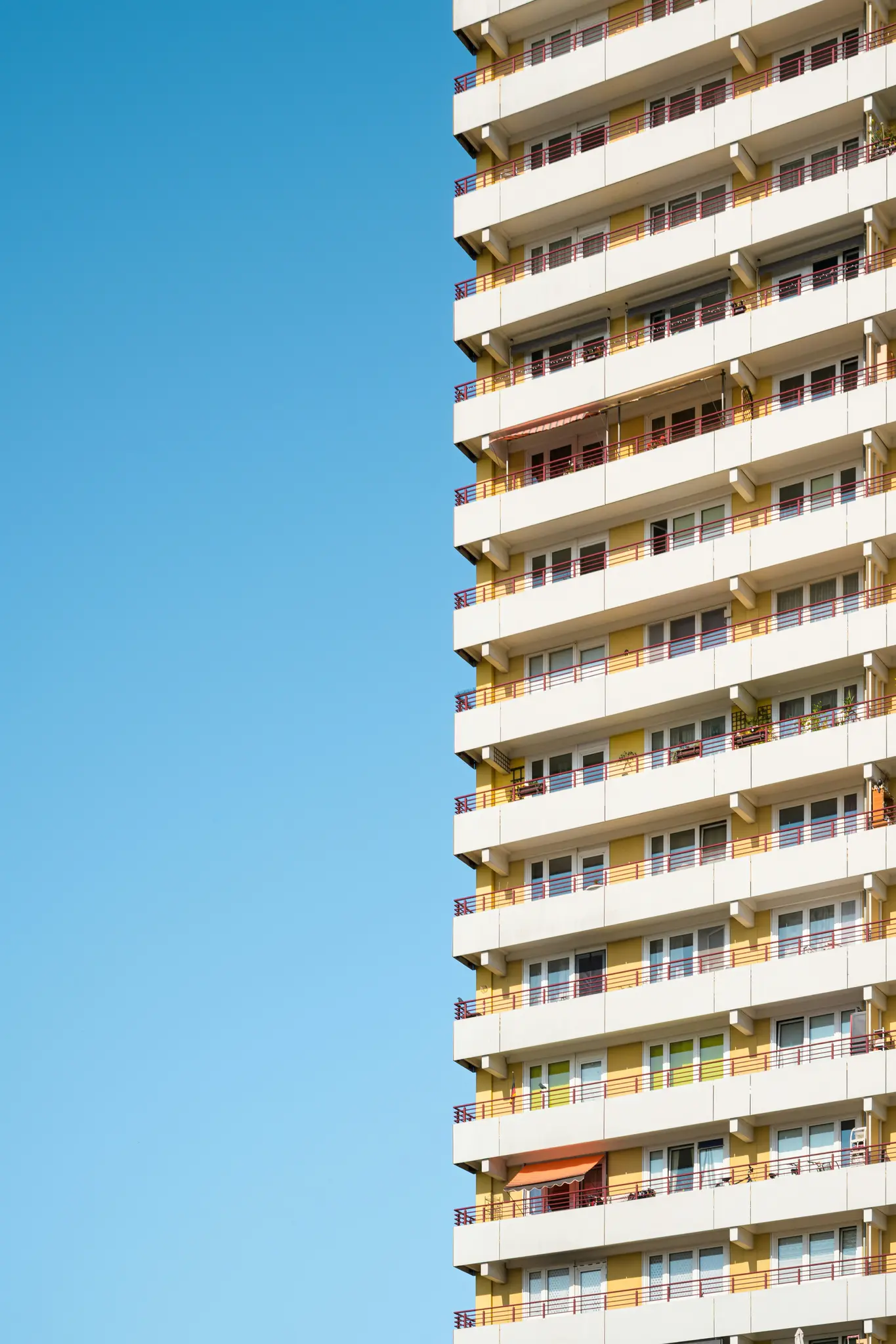 Un edificio de apartamentos alto y amarillo con balcones blancos y barandillas rojas se alza contra un cielo azul brillante y