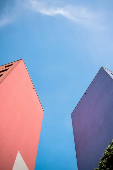 Eine Ansicht von unten auf zwei hohe, flache Gebäudefassaden. Das linke Gebäude ist pink, das rechte violett, beide vor