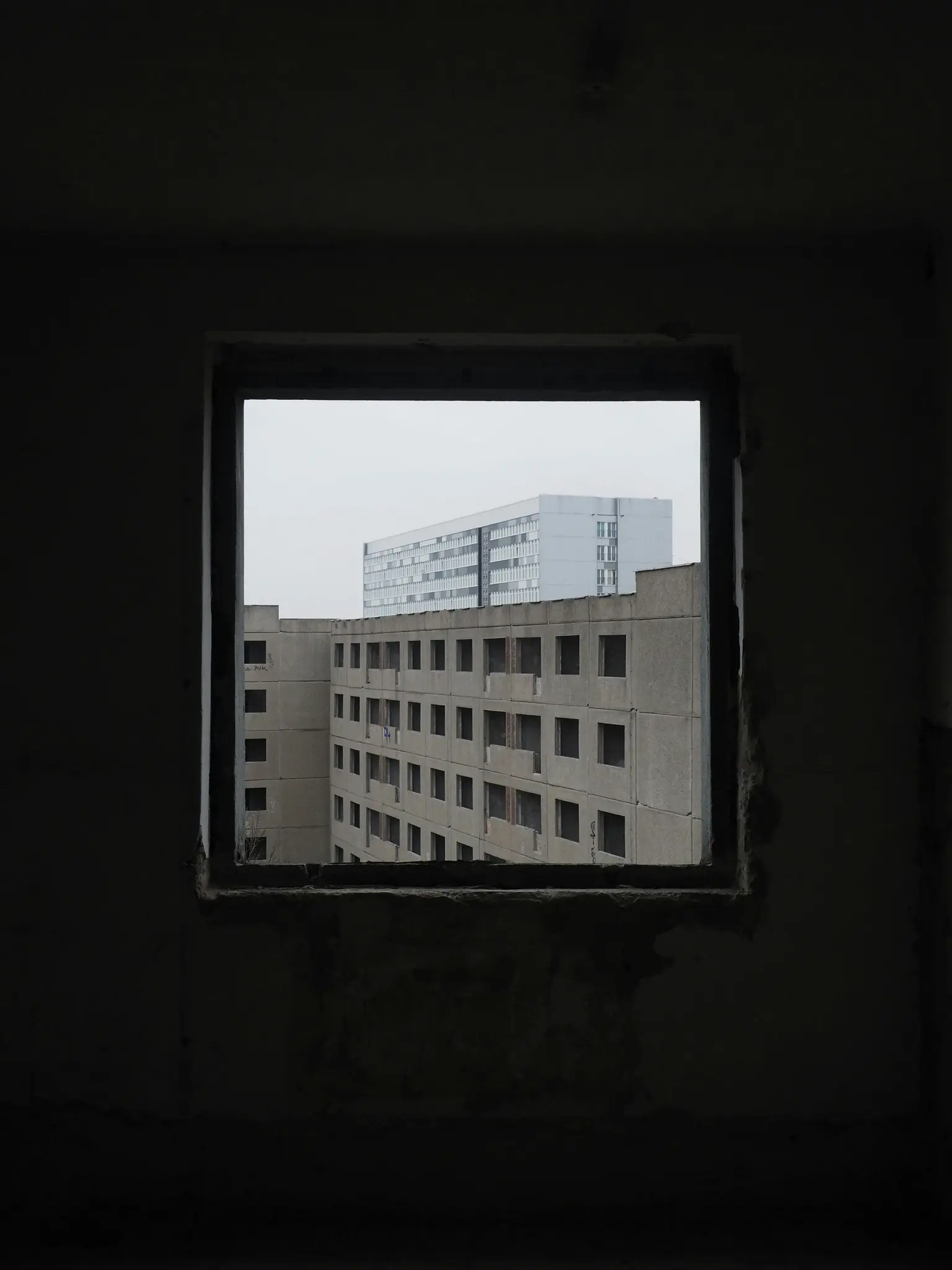 A dark, empty window frame in the foreground looks out onto several concrete buildings under an overcast sky. The interior…
