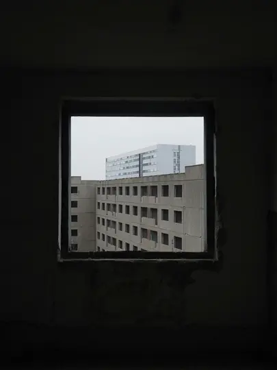 A dark, empty window frame in the foreground looks out onto several concrete buildings under an overcast sky. The interior…