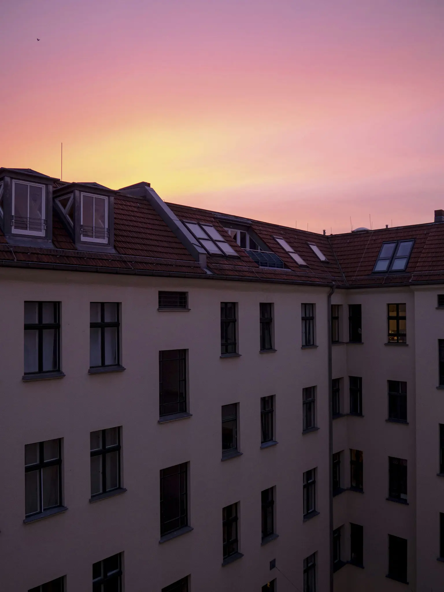 Un edificio con un tejado de tejas rojas y muchas ventanas oscuras se recorta contra un cielo con un degradado de rosa
