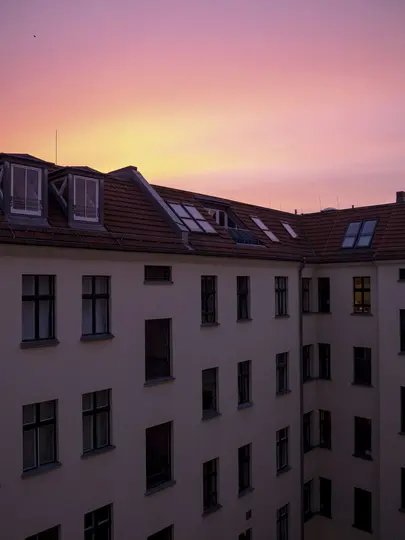 Un edificio con un tetto di tegole rosse e molte finestre scure è in silhouette contro un cielo sfumato di rosa, arancione e