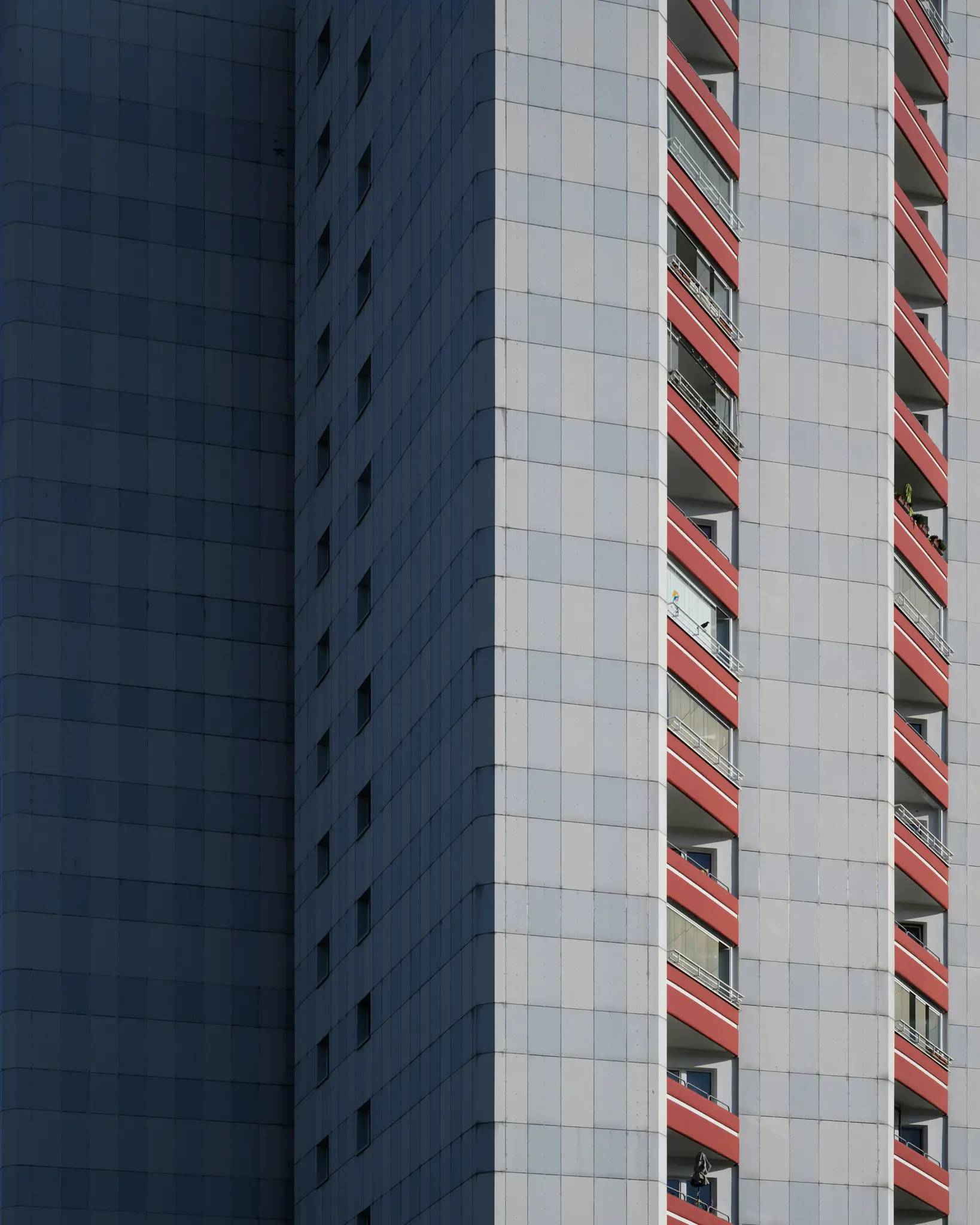 Un edificio alto con fachada de azulejos grises y balcones rojos. El lado izquierdo está en sombra, mientras que el derecho