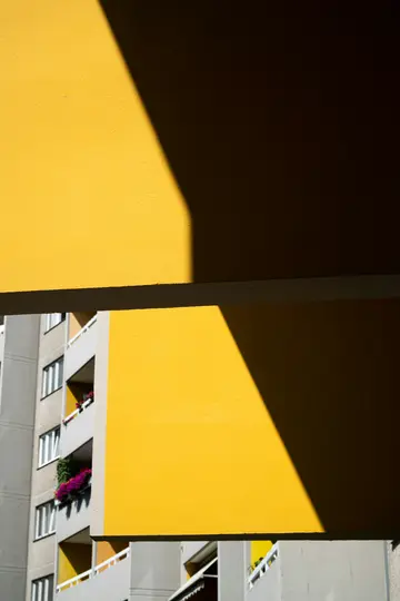 Una pared amarilla vibrante con una sombra diagonal nítida, que contrasta con la fachada gris de un edificio al fondo.