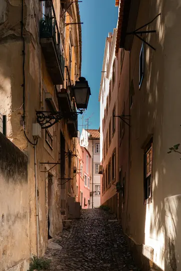 Úzká, do kopce vedoucí dlážděná ulice je lemována vysokými, starými budovami v odstínech béžové, růžové a bílé pod…