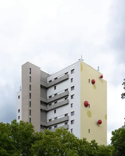 Ein hohes, modernes Wohnhaus unter einem bewölkten Himmel. Eine Seite ist weiß mit grauen Balkonen, die andere ist hellgelb