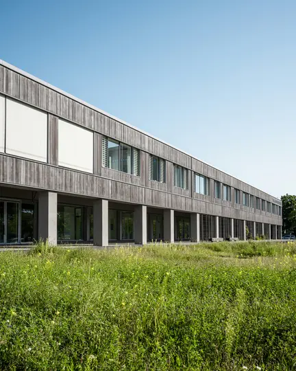 Un edificio moderno con facciata a doghe di legno e molte finestre, alcune con persiane bianche, si trova dietro un campo di