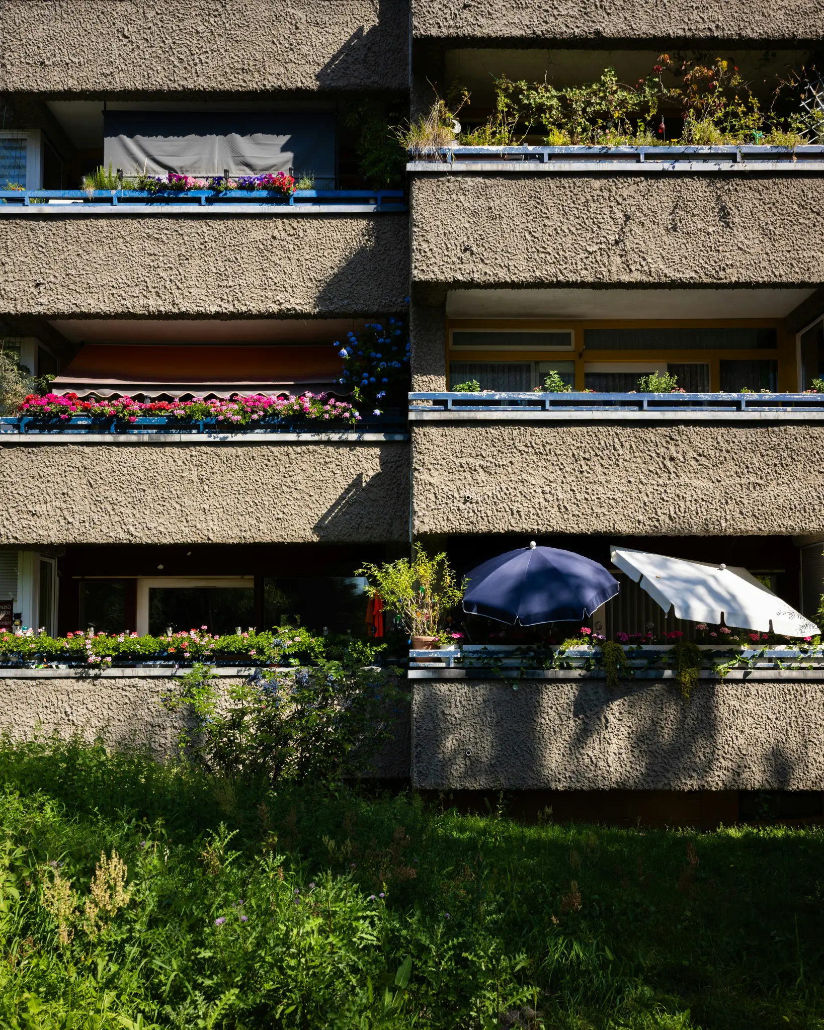 Betónová fasáda budovy s viacerými balkónmi. Každý balkón má kvetináče s farebnými kvetmi a zeleňou...