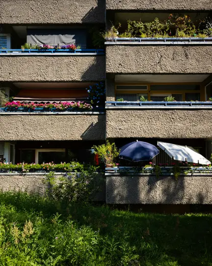 La facciata di un edificio in cemento con più balconi. Ogni balcone presenta fioriere con fiori colorati e vegetazione...