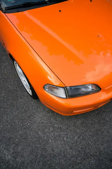 Eine Nahaufnahme von oben eines glänzenden, leuchtend orangefarbenen Autos, das auf dunkelgrauem Asphalt geparkt ist.