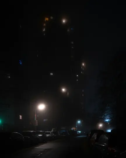 Una escena nocturna oscura y brumosa que muestra una calle flanqueada por coches aparcados y un edificio alto con ventanas