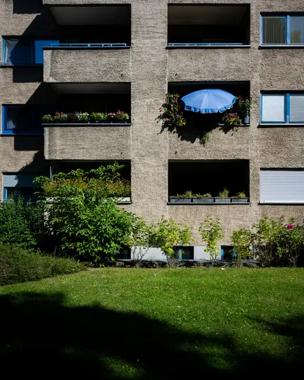 Betonový bytový dům s mnoha balkony a okny. Na jednom z balkonů je otevřený modrý deštník…