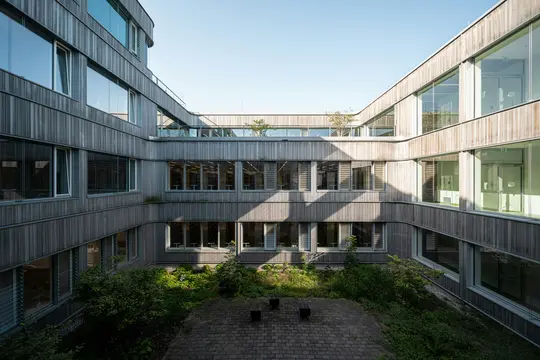 Modernin rakennuksen sisäpiha, jossa vaaleanharmaat puupaneelit ja paljon ikkunoita. Auringonvalo luo voimakkaita varjoja.