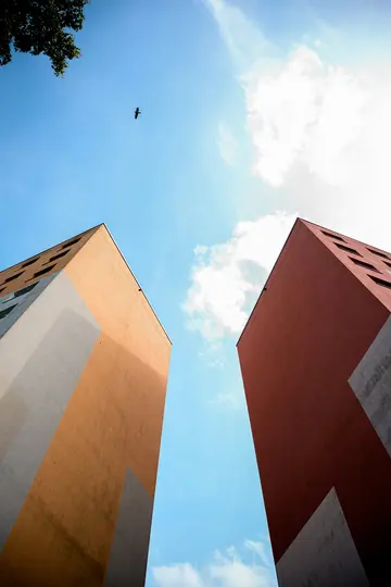 Vista dal basso di due edifici alti e moderni con facciate arancioni e rosse su un cielo blu brillante. Un uccello vola nel…
