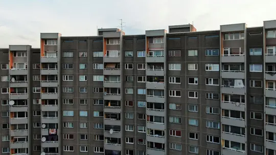 Suuri, harmaa betoninen kerrostalo täyttää kuvan, ja siinä on monia ikkunoita ja parvekkeita pilvisen taivaan alla.