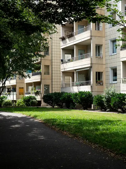 Una vista di un palazzo di colore chiaro con diversi balconi, parzialmente nascosto da alberi e arbusti verde scuro. Un