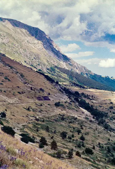 Širokoúhlá fotografie zachycuje rozlehlou horskou krajinu pod zataženou oblohou. Klikatá cesta protíná svahy.
