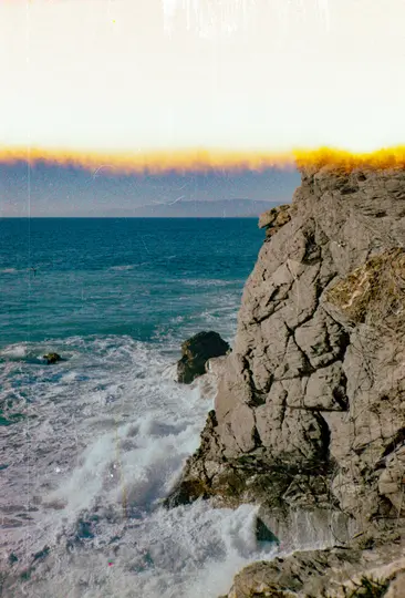 Živá fotografie drsného šedého útesu vpravo, s pěnovými bílými vlnami narážejícími na jeho základnu v hlubokém modrém oceánu.