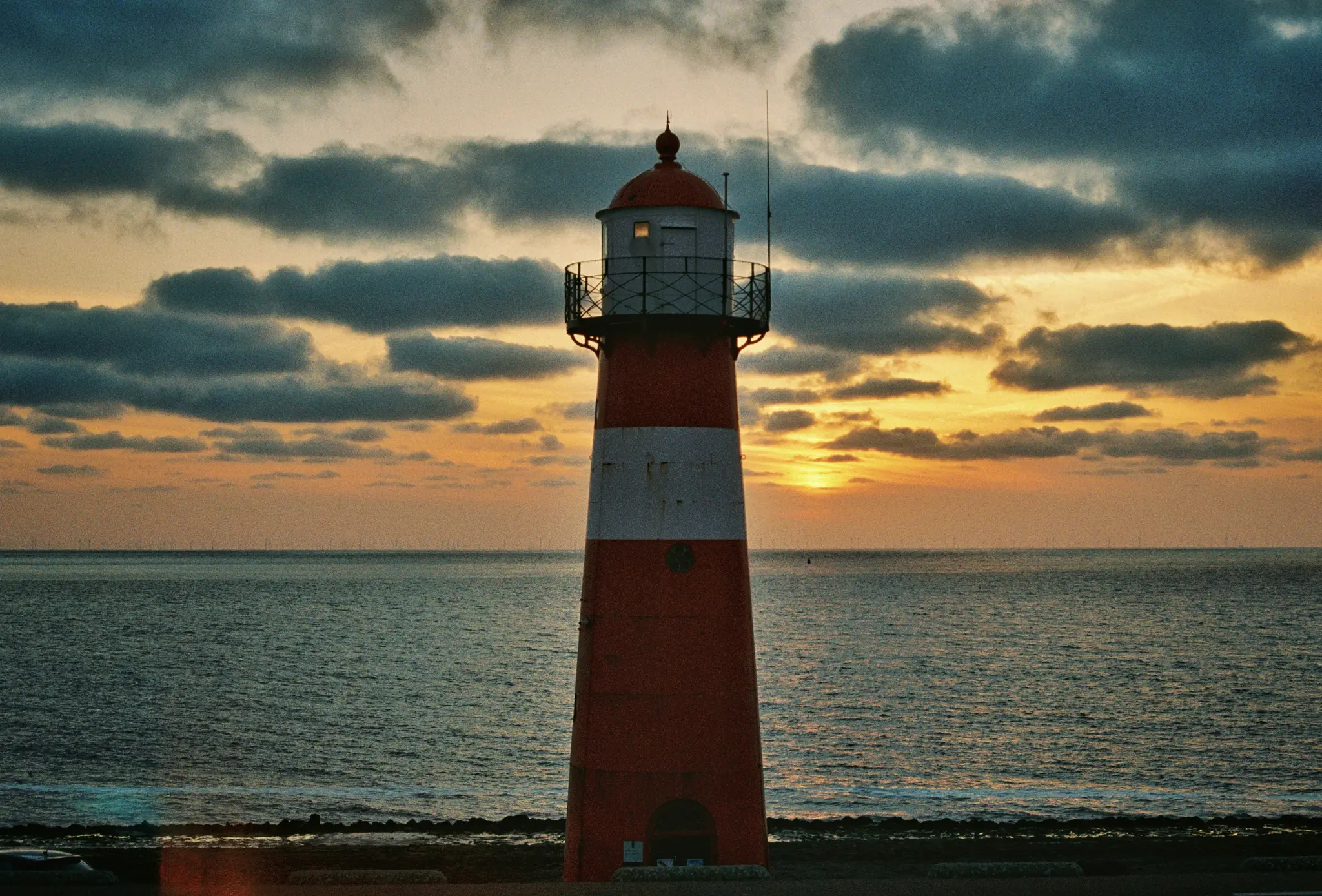 Een hoge rood-wit gestreepte vuurtoren staat in het midden van het frame, gecontrasteerd tegen een levendig oranje en geel…