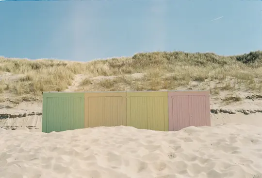Négy pasztellszínű strandfülke (zöld, sárga, sárga, rózsaszín) sora, amelyek részben világos homokba és száraz fűbe vannak