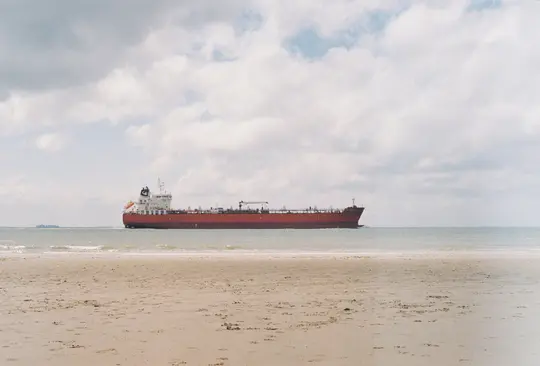 Široký krajinářský záběr ukazuje velkou červenou nákladní loď na oceánu, pohled z písčité pláže. Obloha je zatažená bílými...