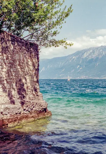 Kamenná zeď se stromem na vrcholu stojí vlevo a vrhá stíny. Velké jezero s modro-zelenou vodou se táhne k…