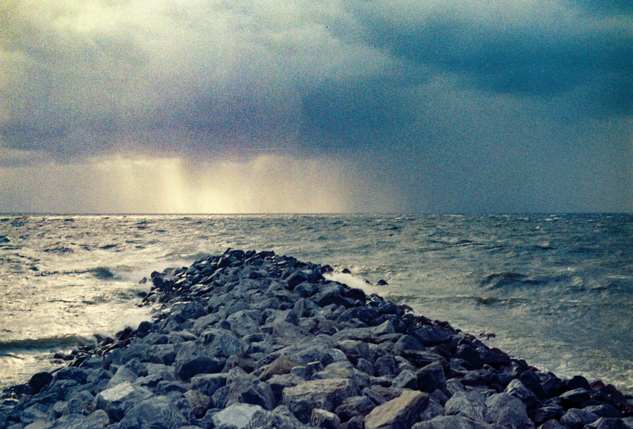 Dlouhé, tmavé skalnaté molo se vine do rozbouřeného, tmavě modrého moře pod těžkým, šedo-modrým bouřkovým oblakem.
