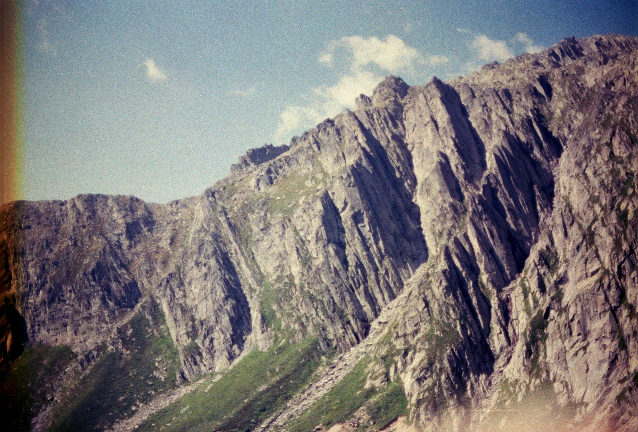 Široká krajinářská fotografie drsného pohoří s prudkými, šedými, skalnatými svahy a plochami zelené trávy. Modrá obloha…