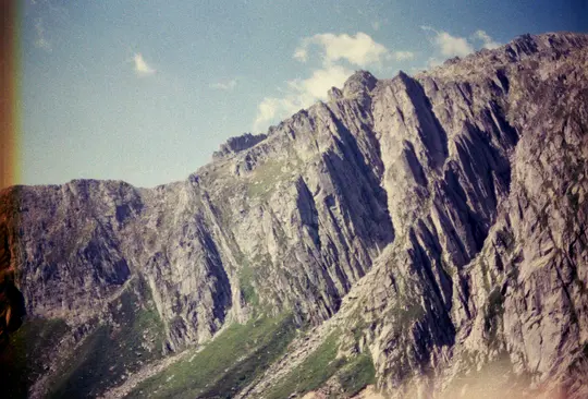 Široká krajinářská fotografie drsného pohoří s prudkými, šedými, skalnatými svahy a plochami zelené trávy. Modrá obloha…