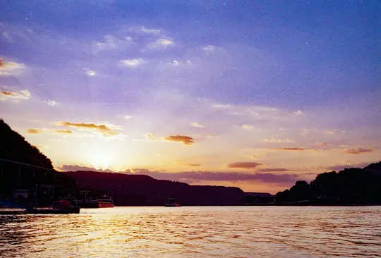Široká krajinářská fotografie řeky při západu slunce. Obloha je fialová a oranžová, slunce zapadá za tmavými kopci.