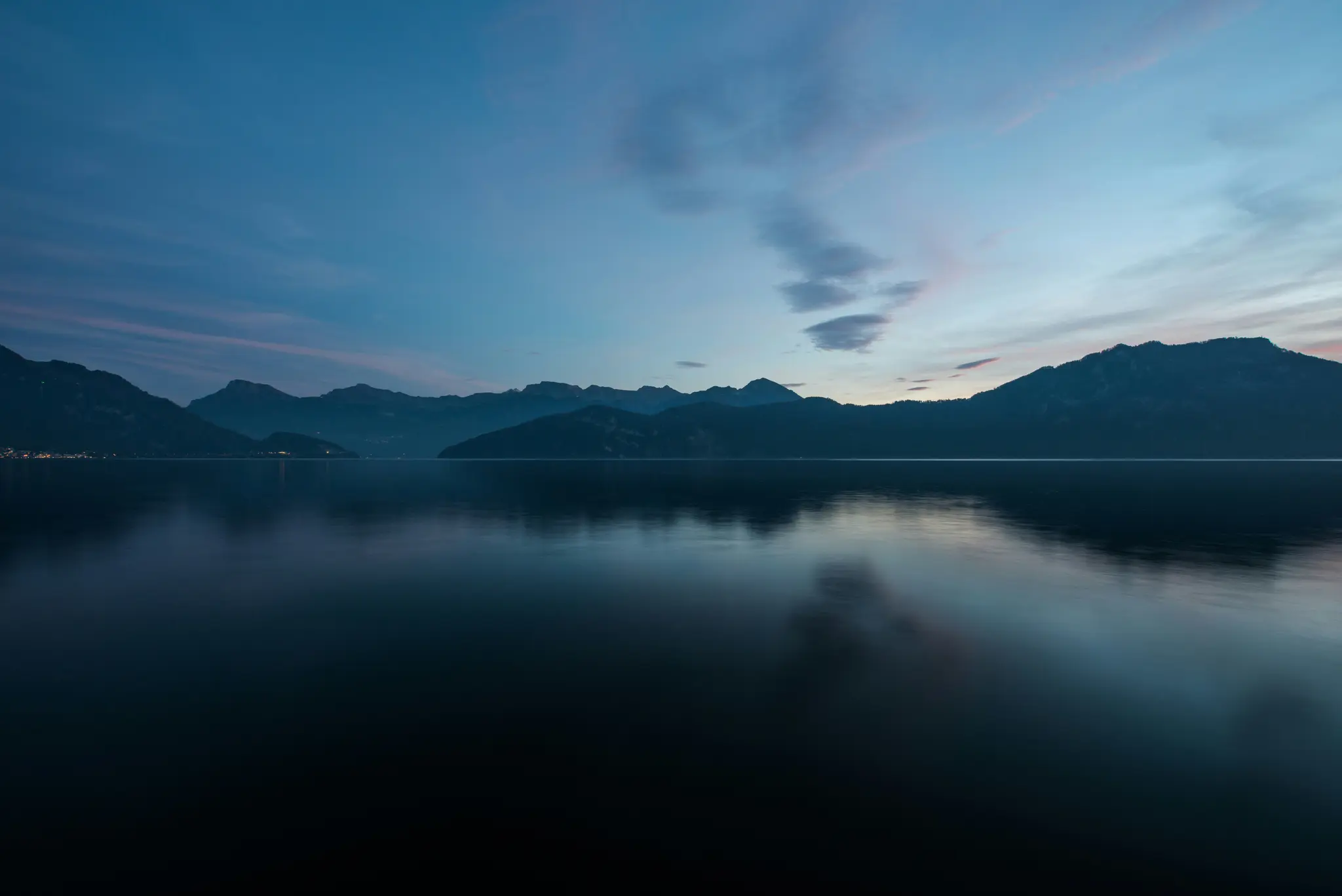 A wide landscape view of a calm lake reflecting the deep blue and pink hues of the twilight sky. Dark mountains line the…