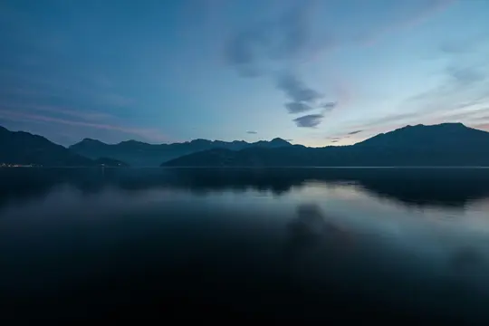 A wide landscape view of a calm lake reflecting the deep blue and pink hues of the twilight sky. Dark mountains line the…