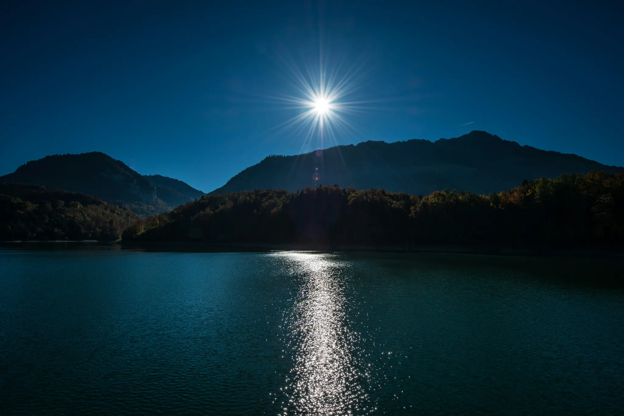 A bright sunburst shines over a dark blue lake, with silhouetted mountains and trees in the background.