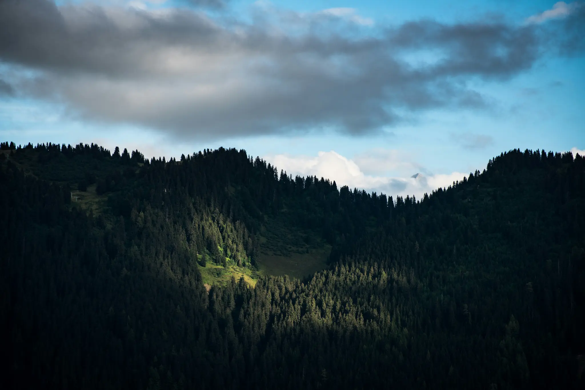 A wide landscape photo of dark green forested mountains under a blue sky with dramatic grey and white clouds. A patch of…