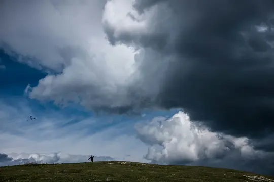 En ensom figur står på en grøn bakke under en dramatisk himmel med mørke stormskyer og blå pletter, en drage flyver til