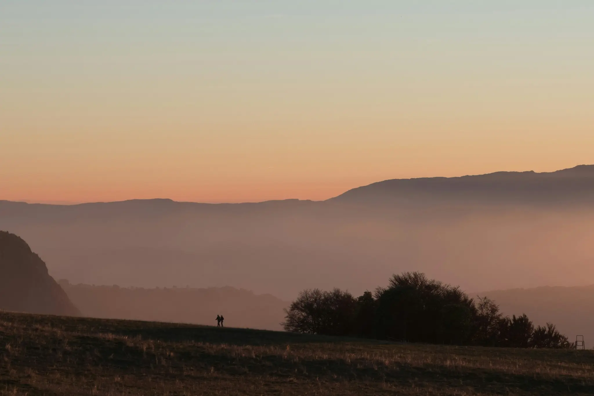 A wide landscape photo showing two silhouetted figures on a grassy hill against a sunset sky with layered mountains.