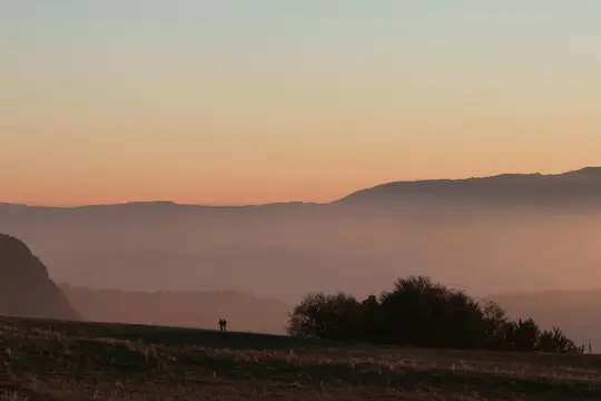 A wide landscape photo showing two silhouetted figures on a grassy hill against a sunset sky with layered mountains.