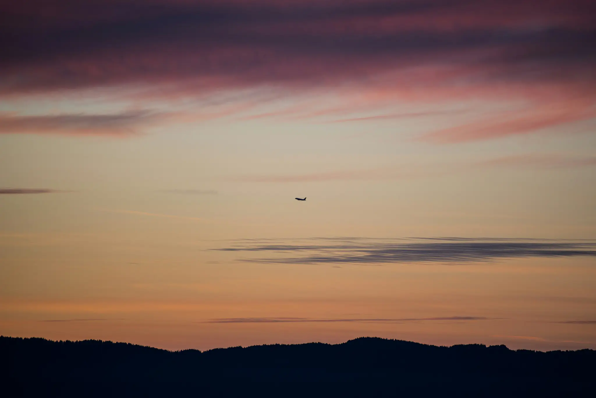 A small, dark silhouette of an airplane flies across a vast sky filled with horizontal clouds in shades of pink, purple…
