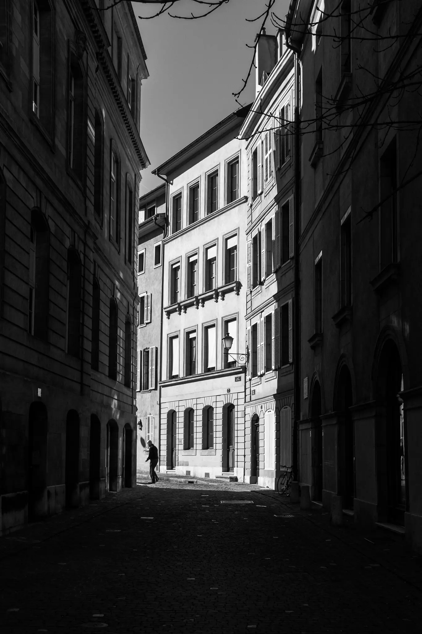 A black and white photo of a narrow, shadowed cobbled street flanked by tall buildings, with a person walking in the…