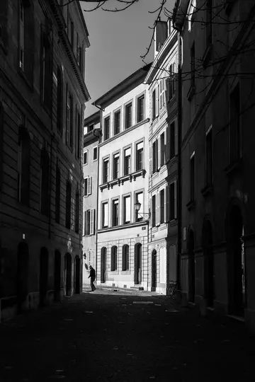 A black and white photo of a narrow, shadowed cobbled street flanked by tall buildings, with a person walking in the…