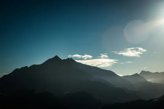 A wide landscape photo of a mountain range silhouetted against a bright blue sky with lens flare and scattered clouds.