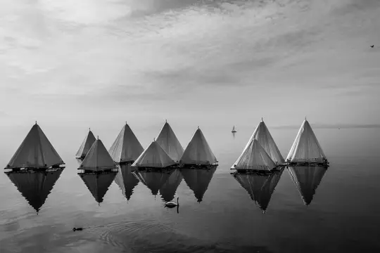 Sort-hvidt billede af flere pyramideformede strukturer, der flyder på roligt vand og reflekteres på overfladen. En lille…