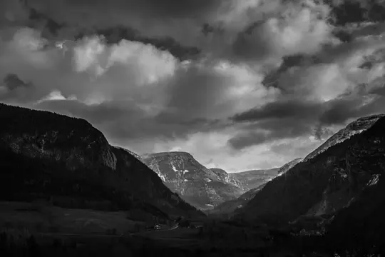 A black and white landscape of a deep mountain valley with dark, forested slopes under a dramatic, cloudy sky. Some distant…