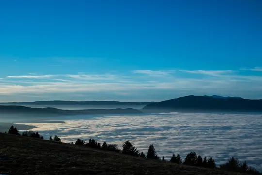 A wide landscape photo shows a valley completely covered by a thick, white layer of clouds, resembling a sea of fog. Dark…