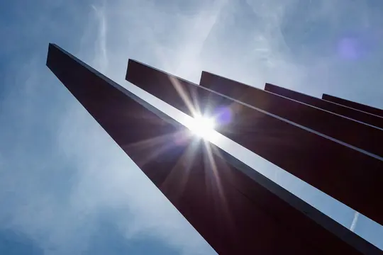 Et lavvinklet billede af solen, der bryder igennem mørke, vinklede arkitektoniske lameller mod en klar blå himmel med tynde