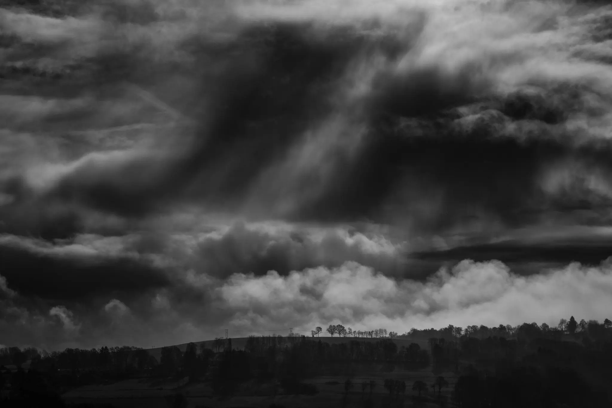 A black and white landscape photo showing a dark, cloudy sky with rays of light breaking through, above a distant treeline…